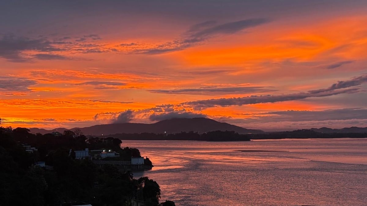 Dramatic sunset over a volcanic island with orange sky reflected in calm water and silhouetted trees on shore