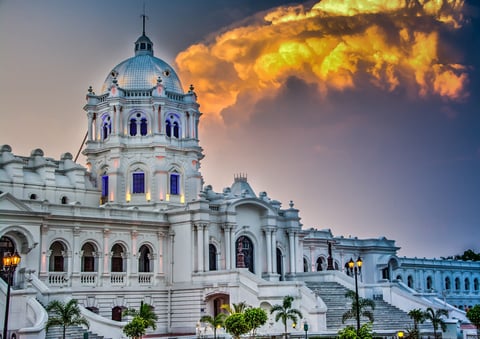 White neoclassical government building with blue dome and tower at sunset, palm trees in foreground, dramatic golden clouds