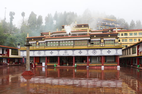 Tibetan Buddhist temple courtyard with ornate red and gold buildings reflected in wet pavement, mountains and mist in background