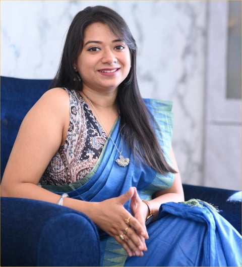 Woman with long black hair smiling, wearing a blue saree and patterned blouse, sitting on a blue sofa
