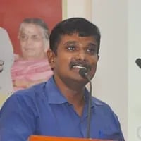 A man with dark hair and mustache wearing a blue polo shirt, speaking at an event with a blurred background