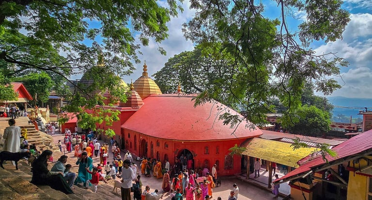 Red domed Hindu temple with golden spire surrounded by green trees, visitors in colorful clothing gathered in the courtyard on a sunny day