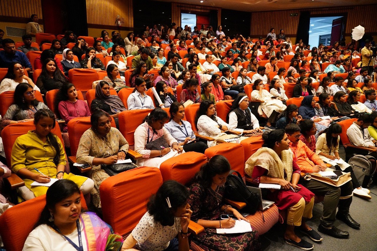 Large audience seated in auditorium with orange chairs, holding papers during a presentation or event