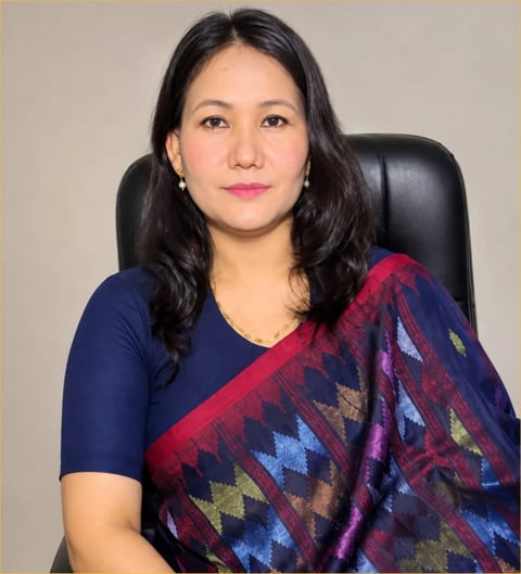 Professional headshot of an Asian woman with long black hair wearing a navy blue top and colorful patterned shawl, seated in a black chair against a neutral background