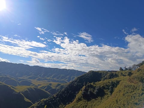 Scenic mountain valley landscape with rolling green hills, forested slopes, and blue sky with white clouds