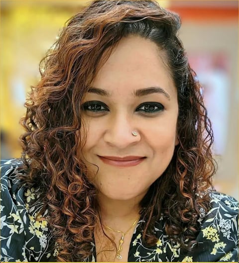 Woman with long curly reddish-brown hair wearing patterned dark top with floral and star design, smiling at camera