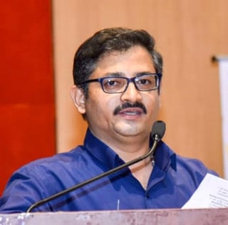 Man wearing blue shirt and glasses speaking at microphone during event