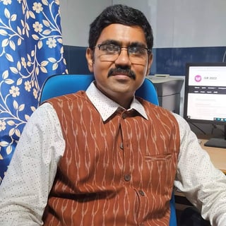 Man with glasses wearing a burgundy pinstriped vest and white shirt, seated at an office desk with blue chair