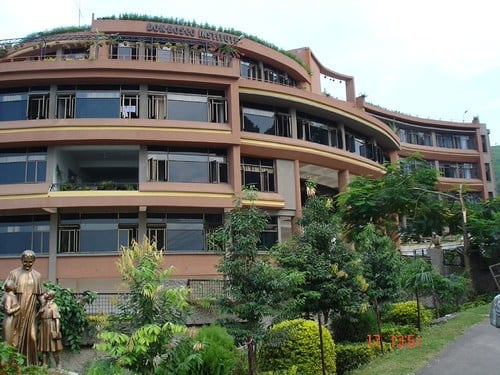 Modern brown residential building with curved architecture, multiple levels, large windows, and landscaped gardens with trees in front