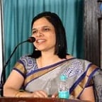 Woman speaking at microphone wearing a traditional patterned saree with a blue wall background