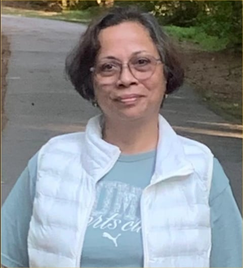 Middle-aged woman with short dark hair and glasses wearing a white puffy vest over a light blue shirt, smiling outdoors.