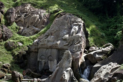 Ancient stone elephant sculpture with ornate carvings nestled among moss-covered rocks and lush green foliage in a forest setting