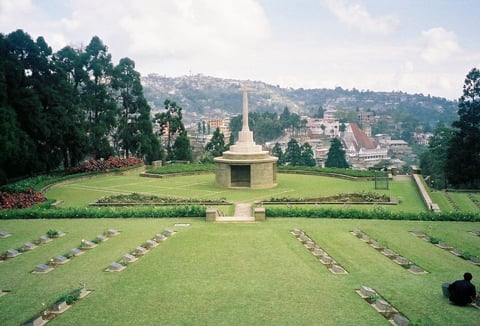 Military cemetery with golden Buddhist stupa in center, rows of graves on green lawn, hills and temples visible in background