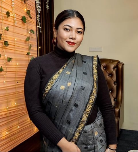 Woman in gray sari with gold trim and black long-sleeve top, seated on leather chair, smiling at camera with indoor plant wall backdrop